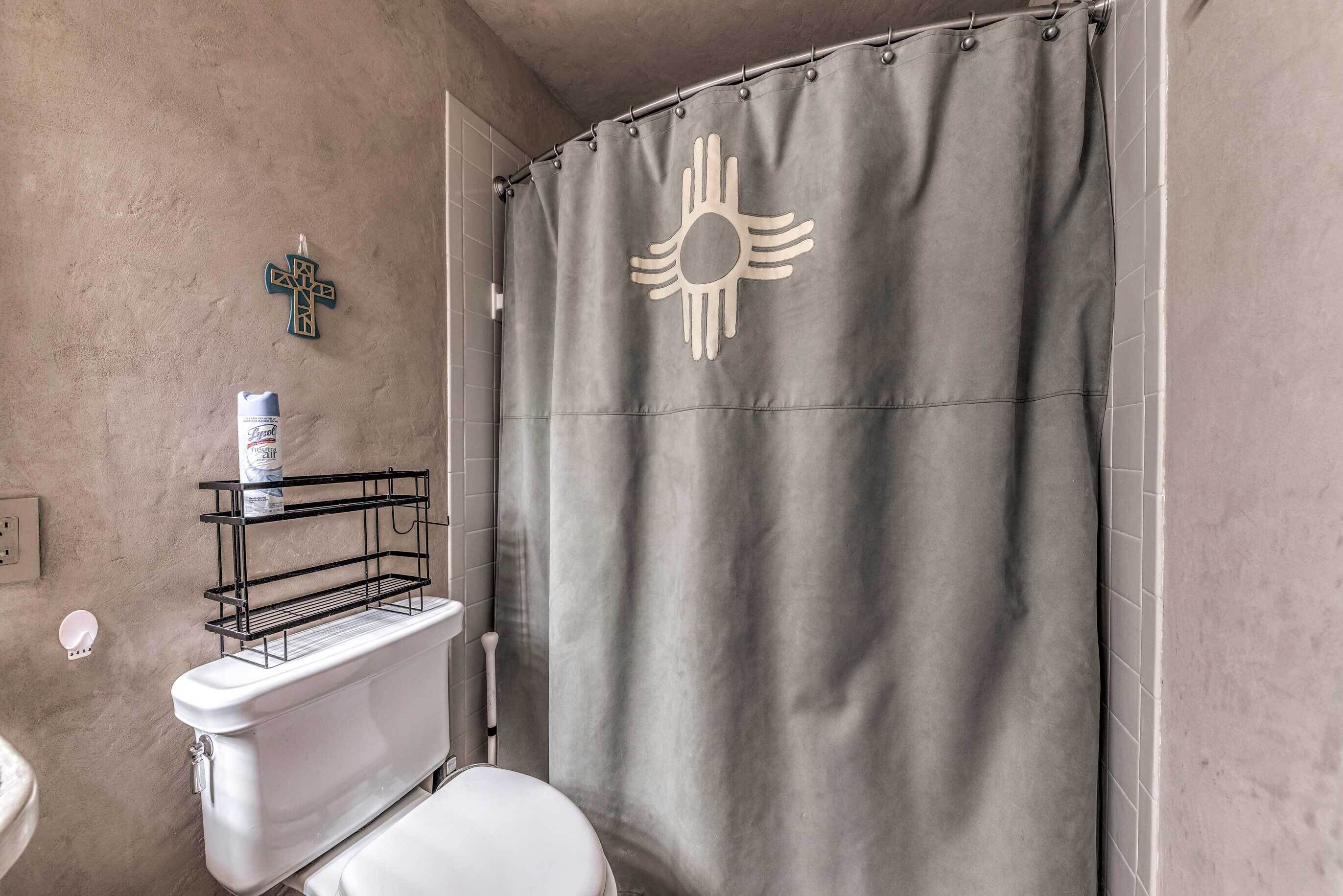 A bathroom with a white toilet, a shower curtain with a sun emblem, a metal shelf above the toilet, and a wall-mounted light fixture.