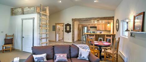 A spacious living area with a brown sofa, a dining table with chairs, a kitchen in the background, and a ceiling fan.