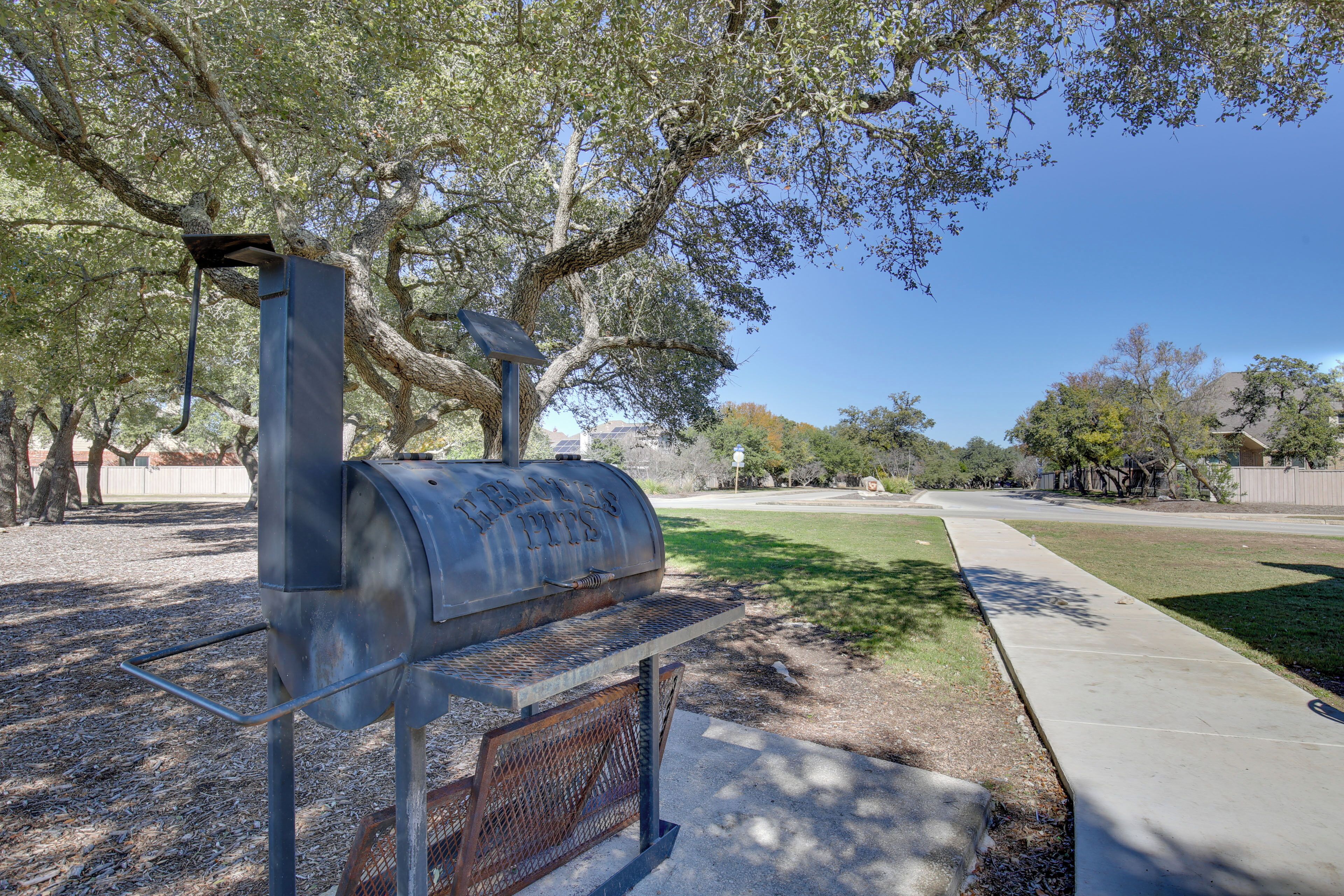 A barbecue grill with a metal enclosure and a chimney, situated in a park with trees and a sidewalk.