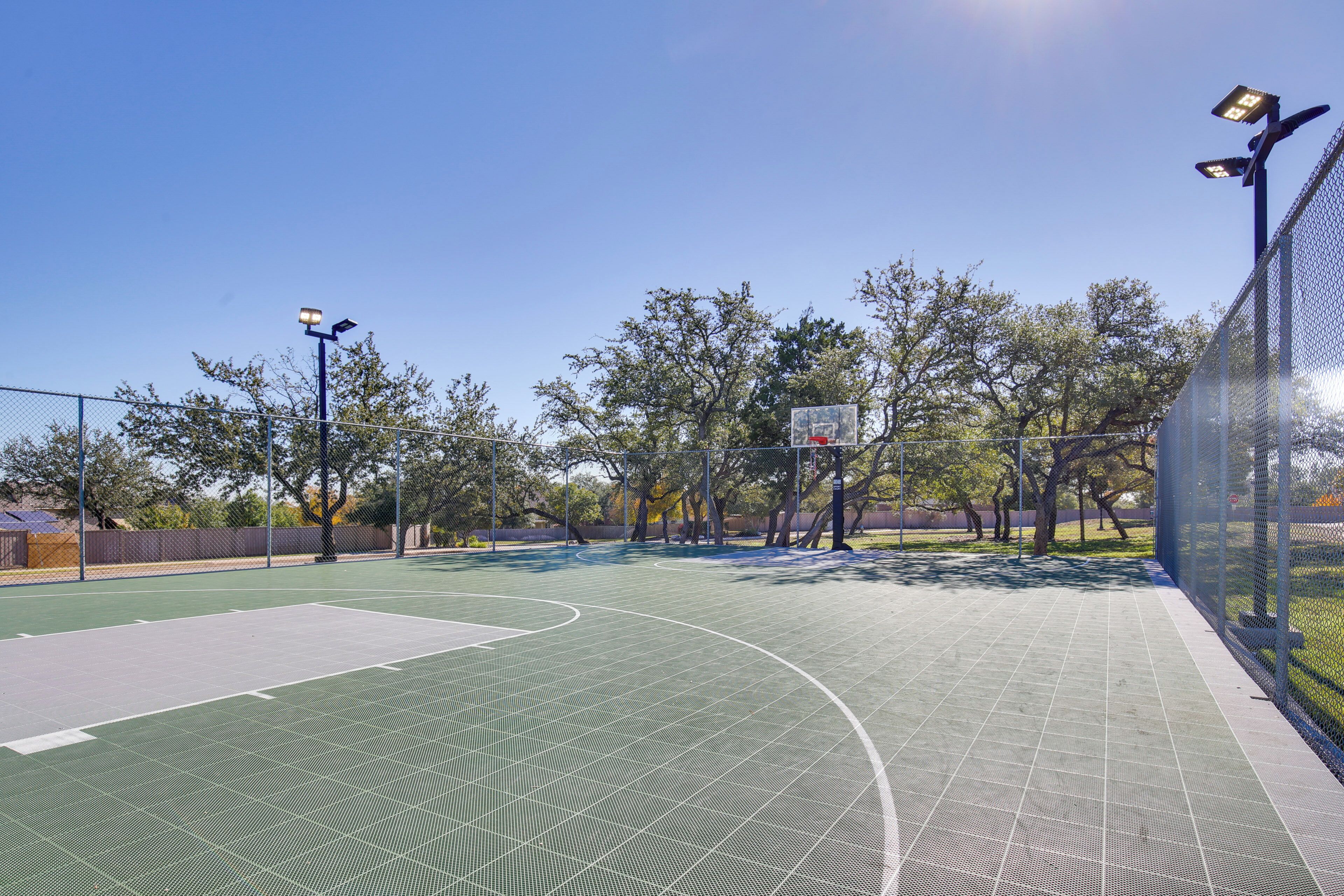 Ein Basketballplatz im Freien mit grüner Spielfläche, umgeben von einem Maschendrahtzaun und mit Beleuchtung ausgestattet.
