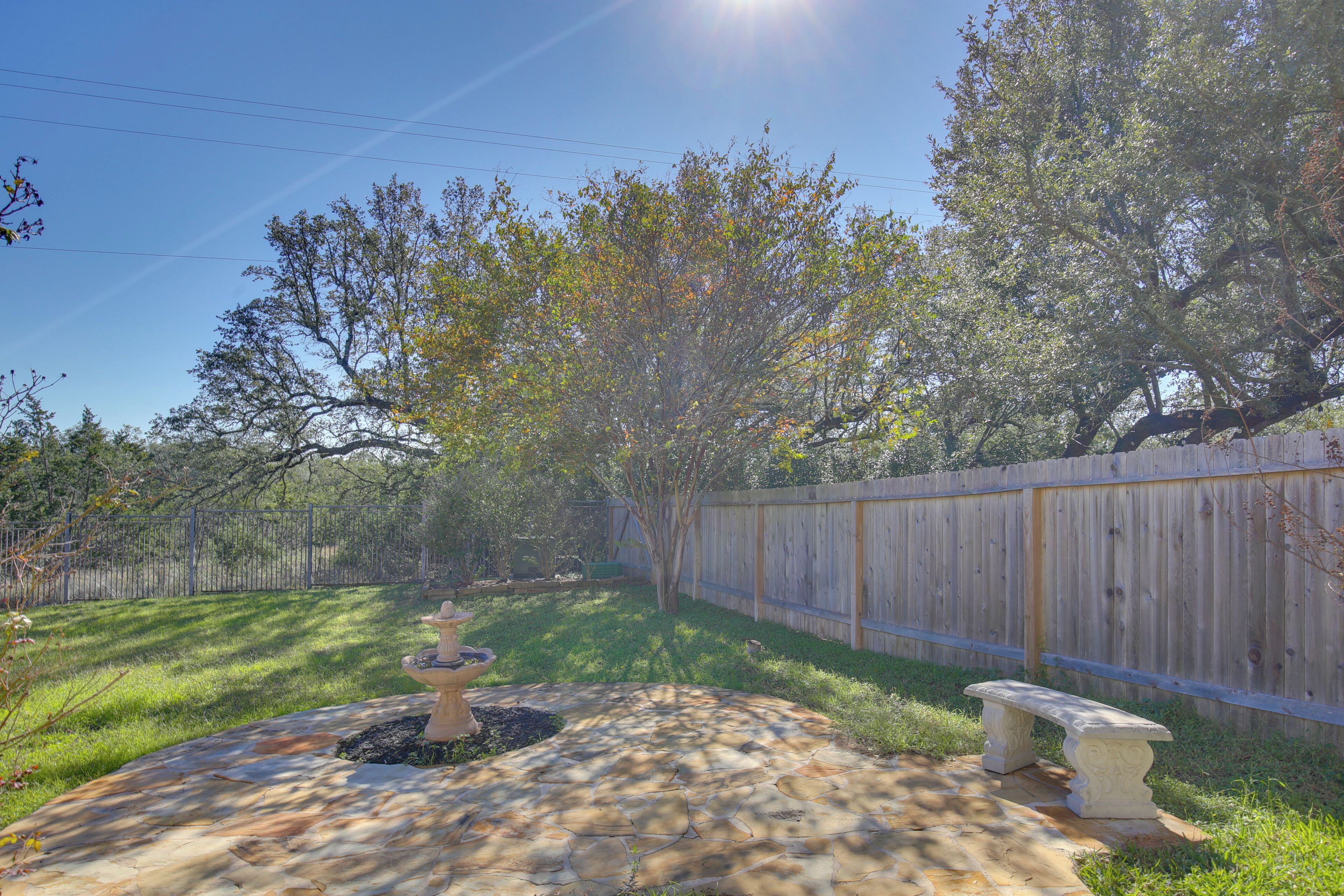 A stone patio with a fountain, a bench, and a wooden fence in a backyard.
