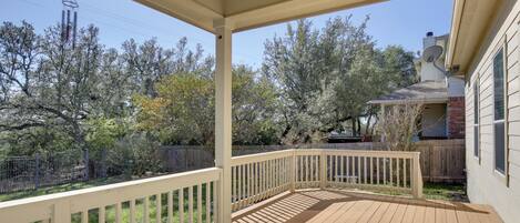 A covered outdoor deck with a wooden floor and white railings, surrounded by trees and a wooden fence.