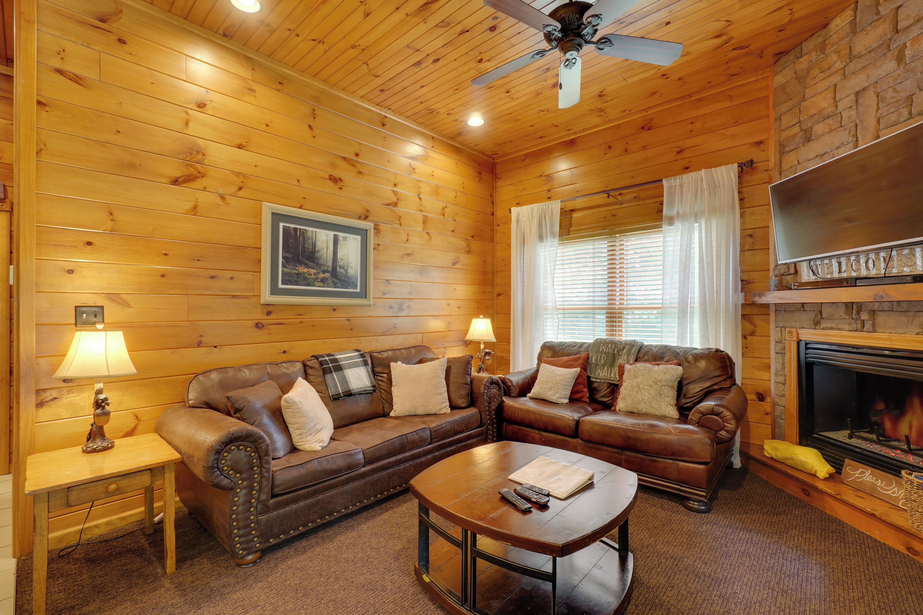 A cozy cabin interior with wooden walls, a fireplace, a leather sofa, a coffee table, and a ceiling fan.