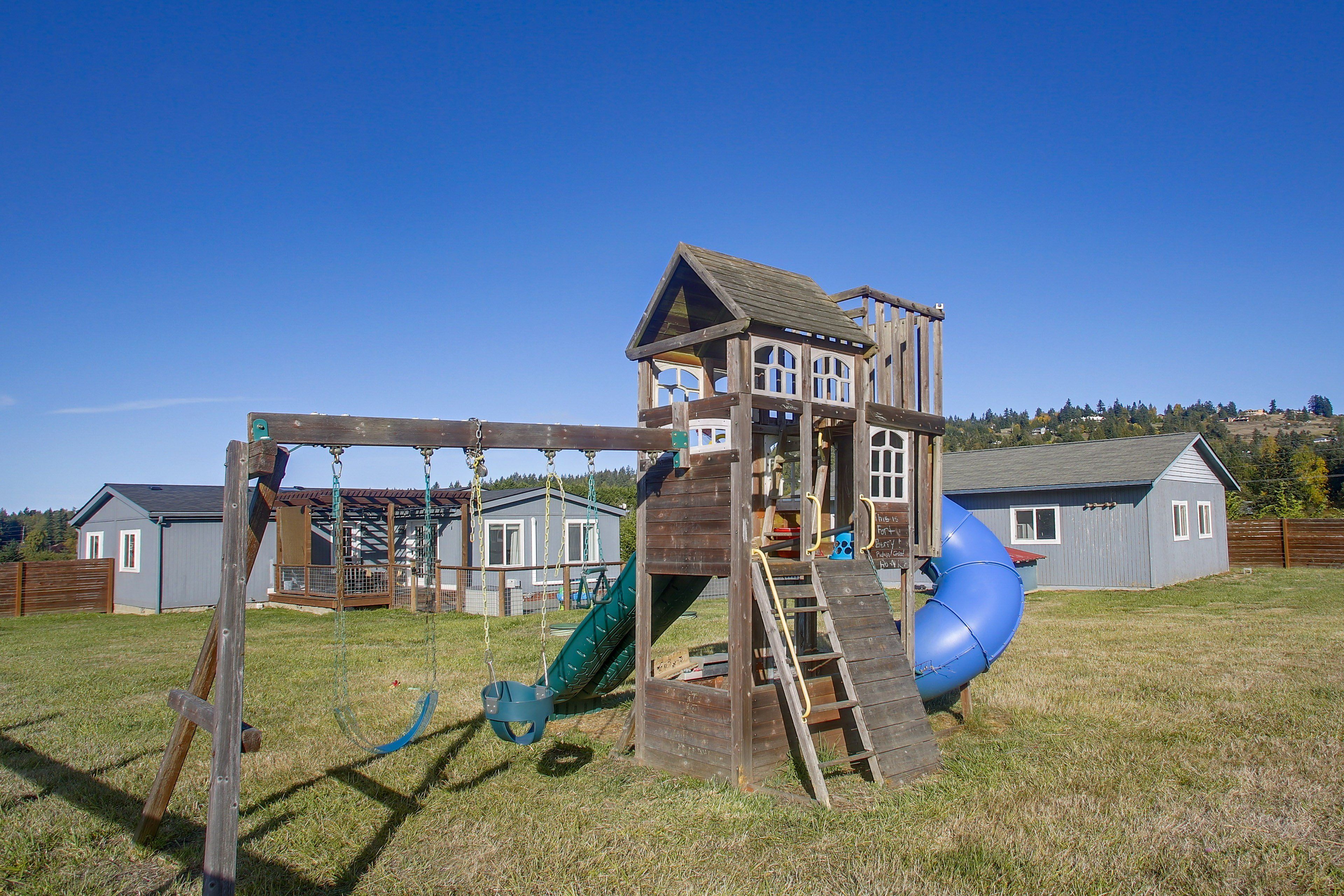 A wooden play structure with a slide and climbing elements, surrounded by a grassy yard and residential buildings.