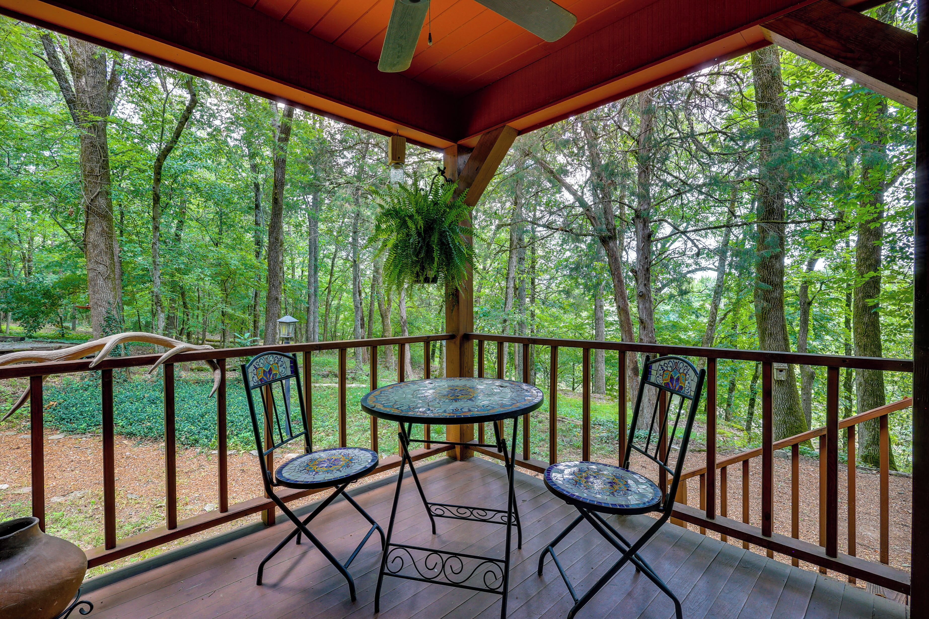 A covered outdoor deck with two chairs and a table, surrounded by a wooden railing, overlooking a forested area.