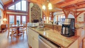 A spacious kitchen with a stone fireplace, wooden beams, and a large island with a coffee maker. - Spacious Virginia Vacation Rental w/ Yard & Sauna (Purcellville)