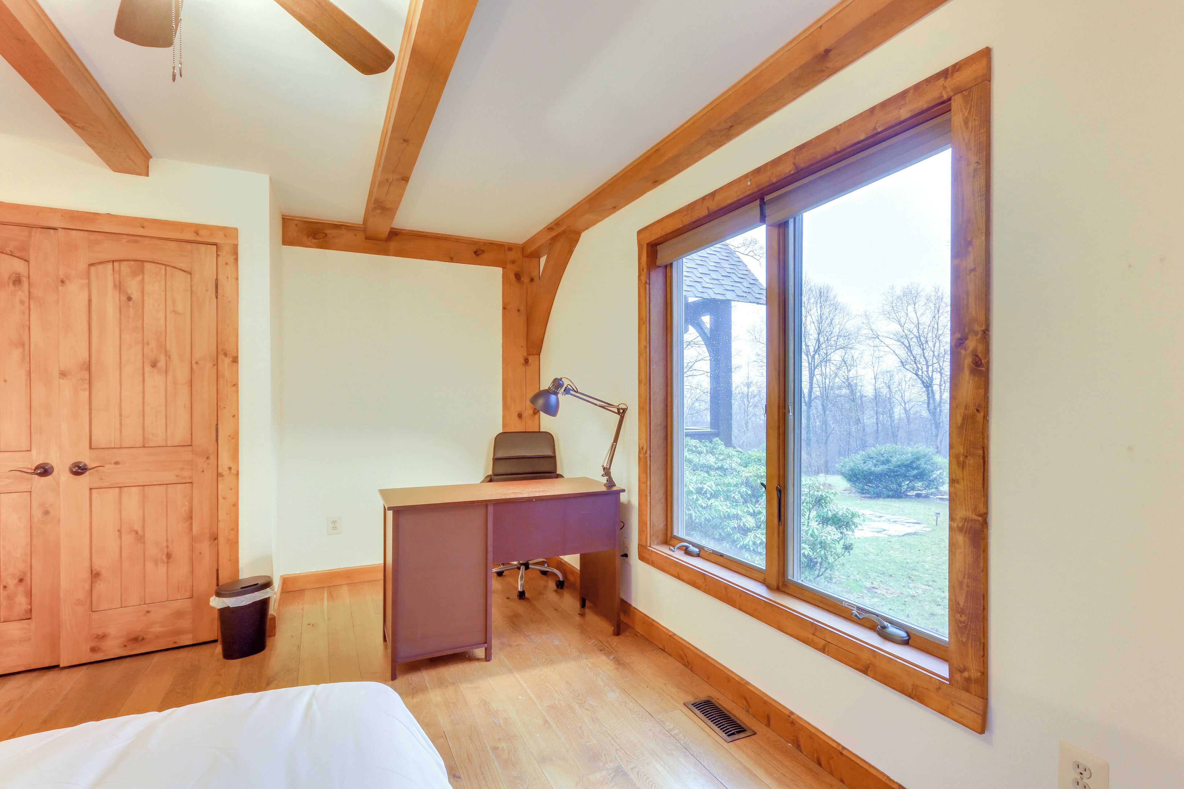 A bedroom with a wooden ceiling, a large window, a desk with a lamp, and a bed.