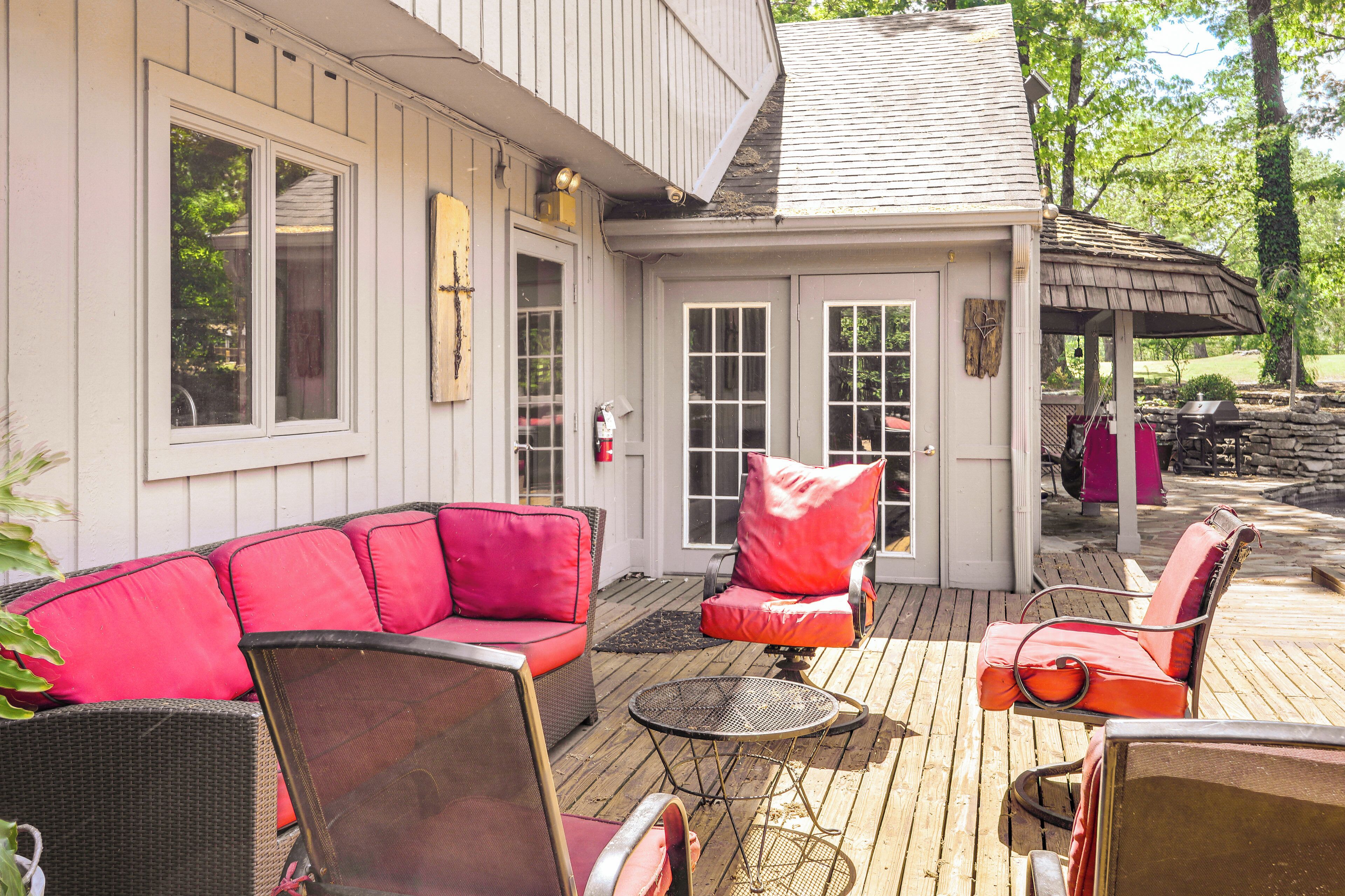 A covered outdoor seating area with wicker furniture and red cushions, a wooden deck, and a stone wall in the background.