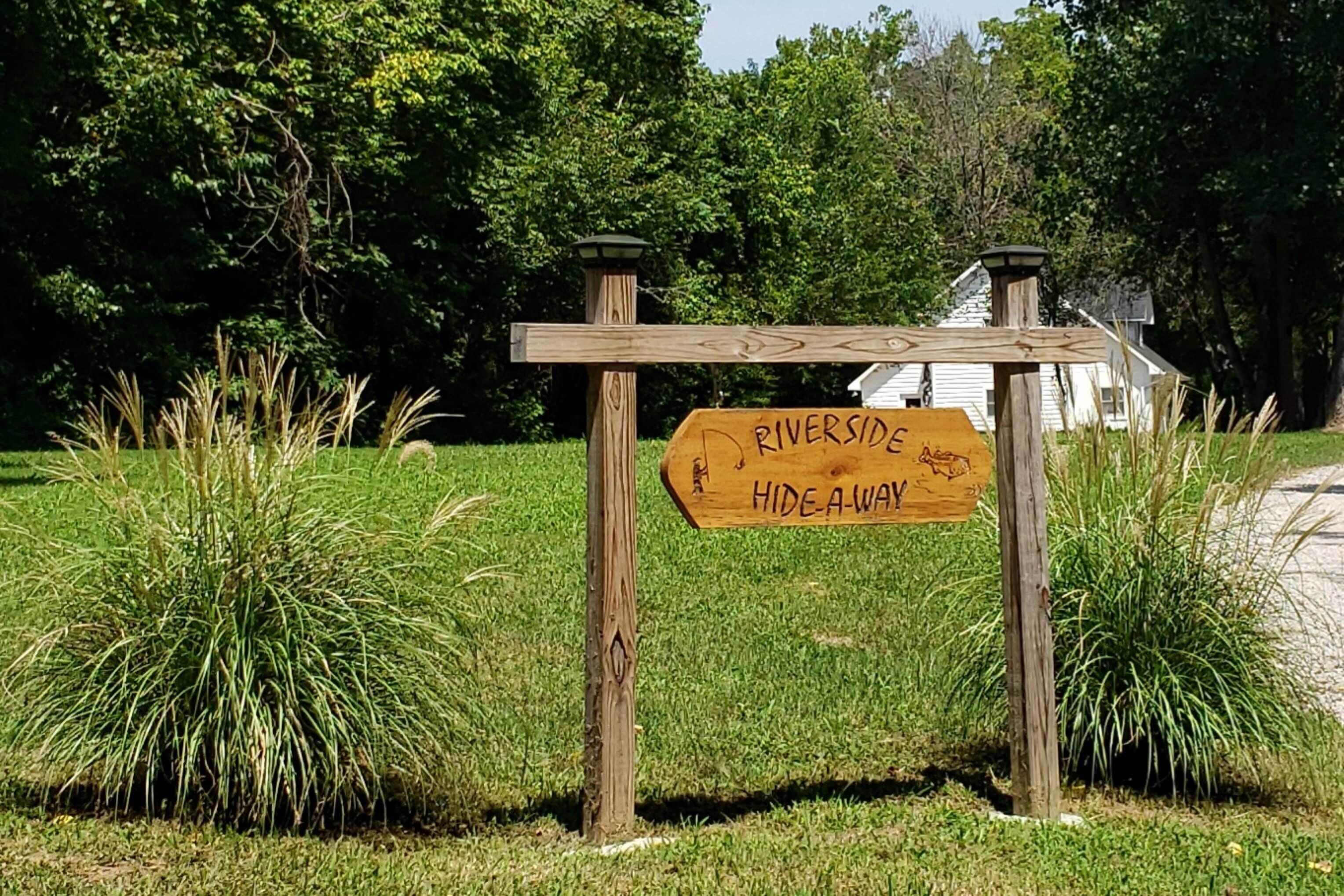 Un senyal de fusta amb la inscripció 'Riverside Hide-A-Way', envoltat d'herba i arbres.