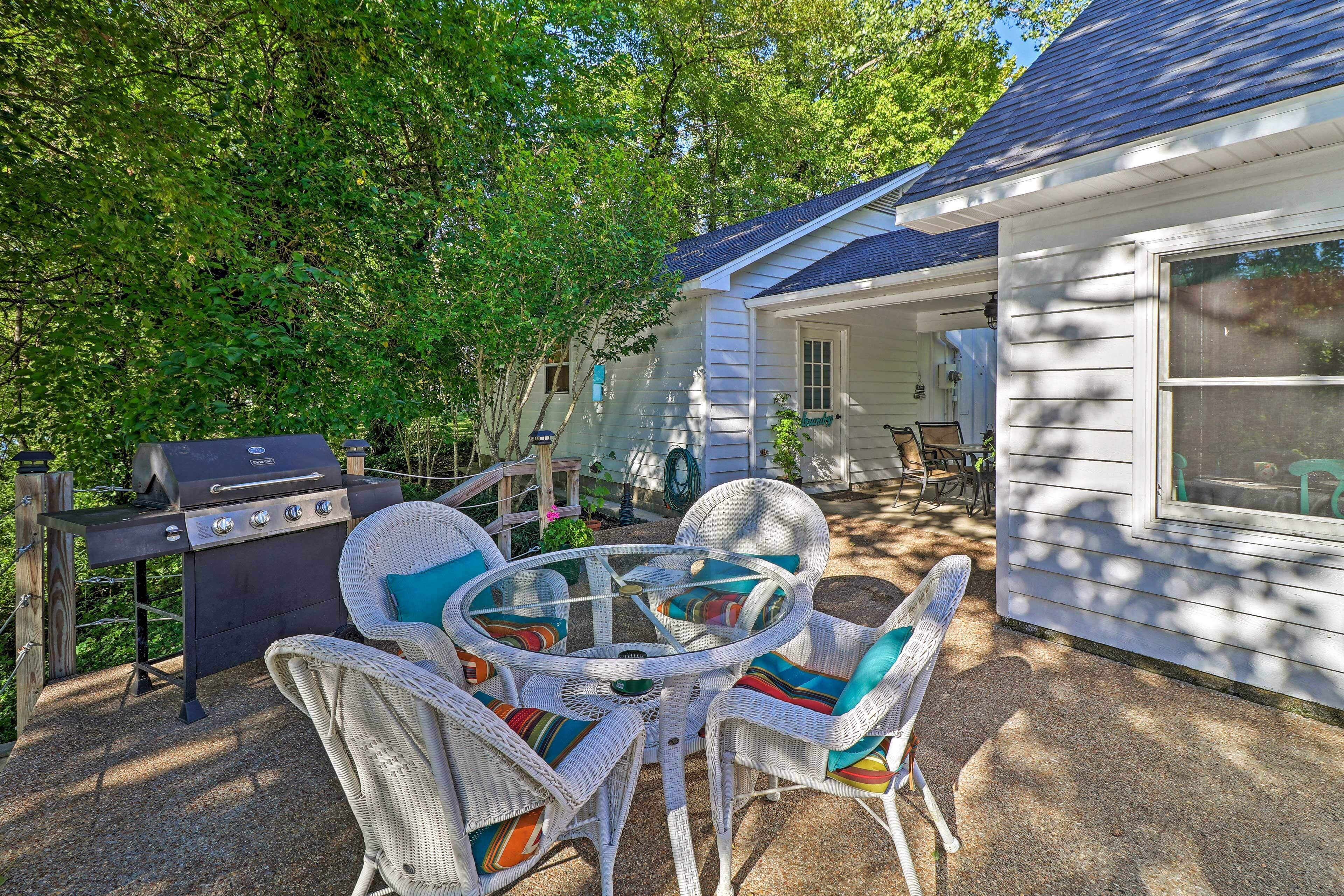 Une terrasse avec des chaises en osier et un barbecue, entourée d’une maison dotée d’un porche abrité.
