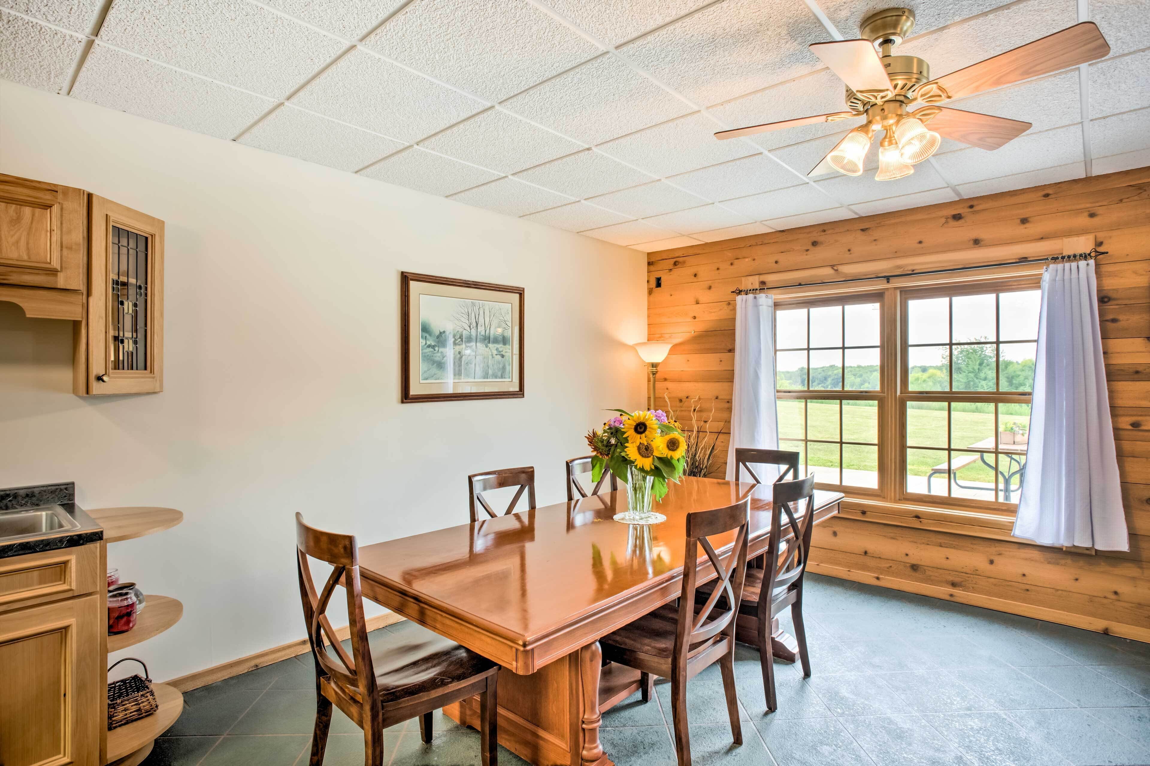 A dining area with a wooden table, chairs, a vase with flowers, a ceiling fan, and a window with curtains.