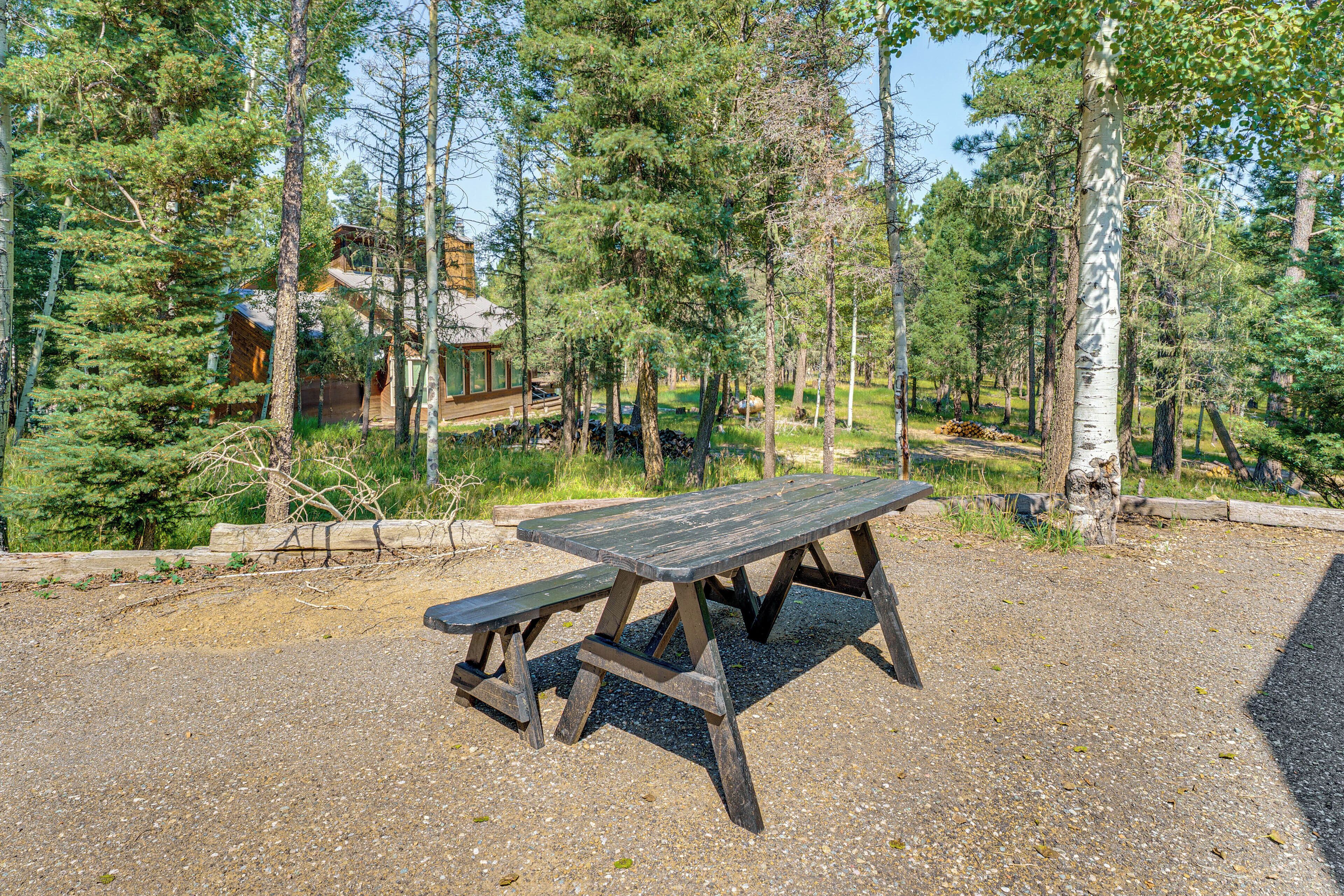 Une table de pique-nique en bois, située dans une zone boisée, avec un chalet en arrière-plan.