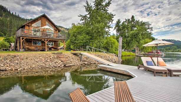 A wooden cabin with a balcony overlooking a lake, a dock with lounge chairs, and a stone pathway leading to the cabin. - Waterfront Cabin w/ 2 Boat Docks & Mtn Views! (St. Maries)