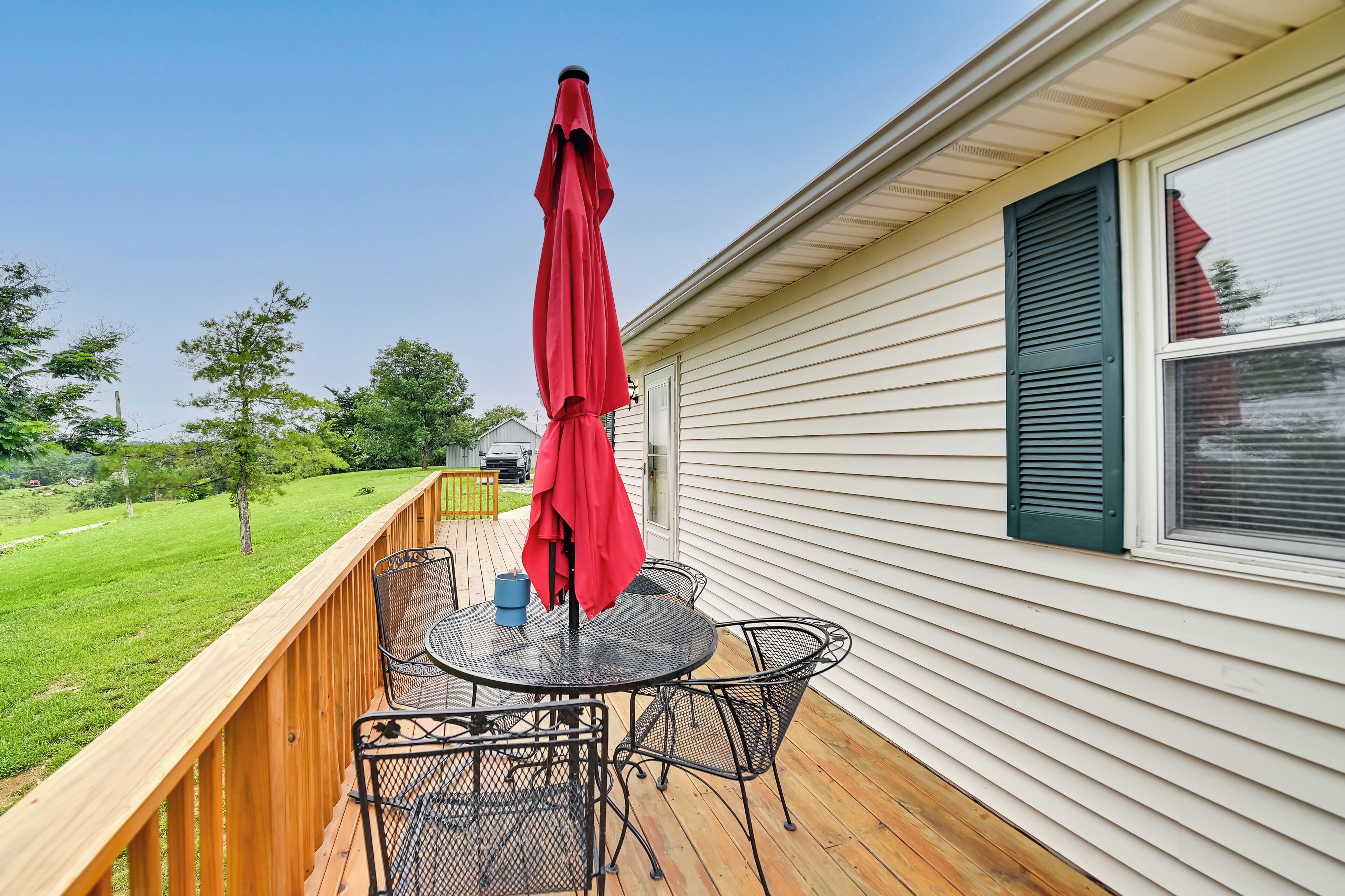 A deck with a round table and chairs, a red umbrella, and a view of a grassy yard.
