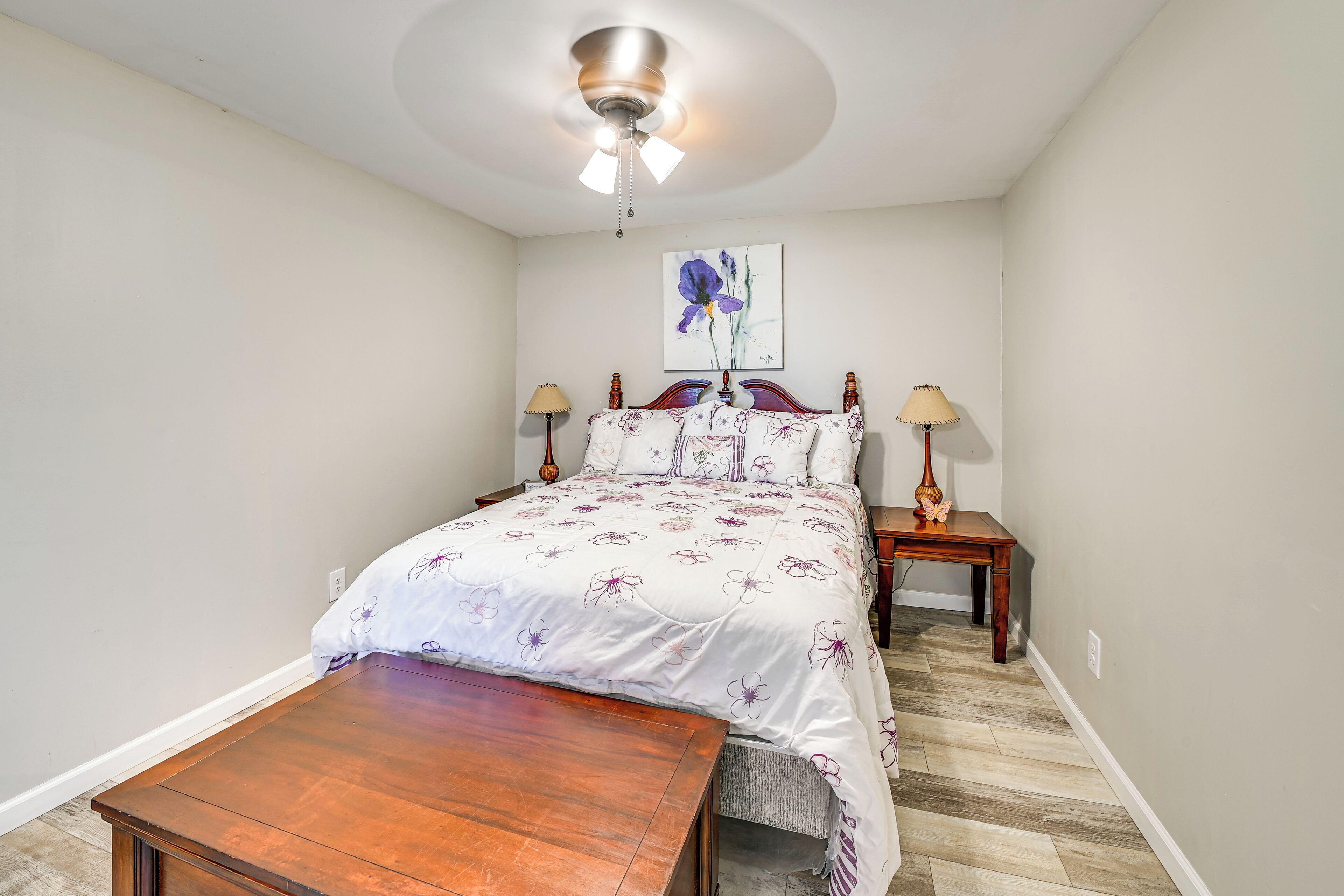 Une chambre à coucher avec un lit en bois, une table de chevet, une lampe, un ventilateur de plafond et un tableau accroché au mur.