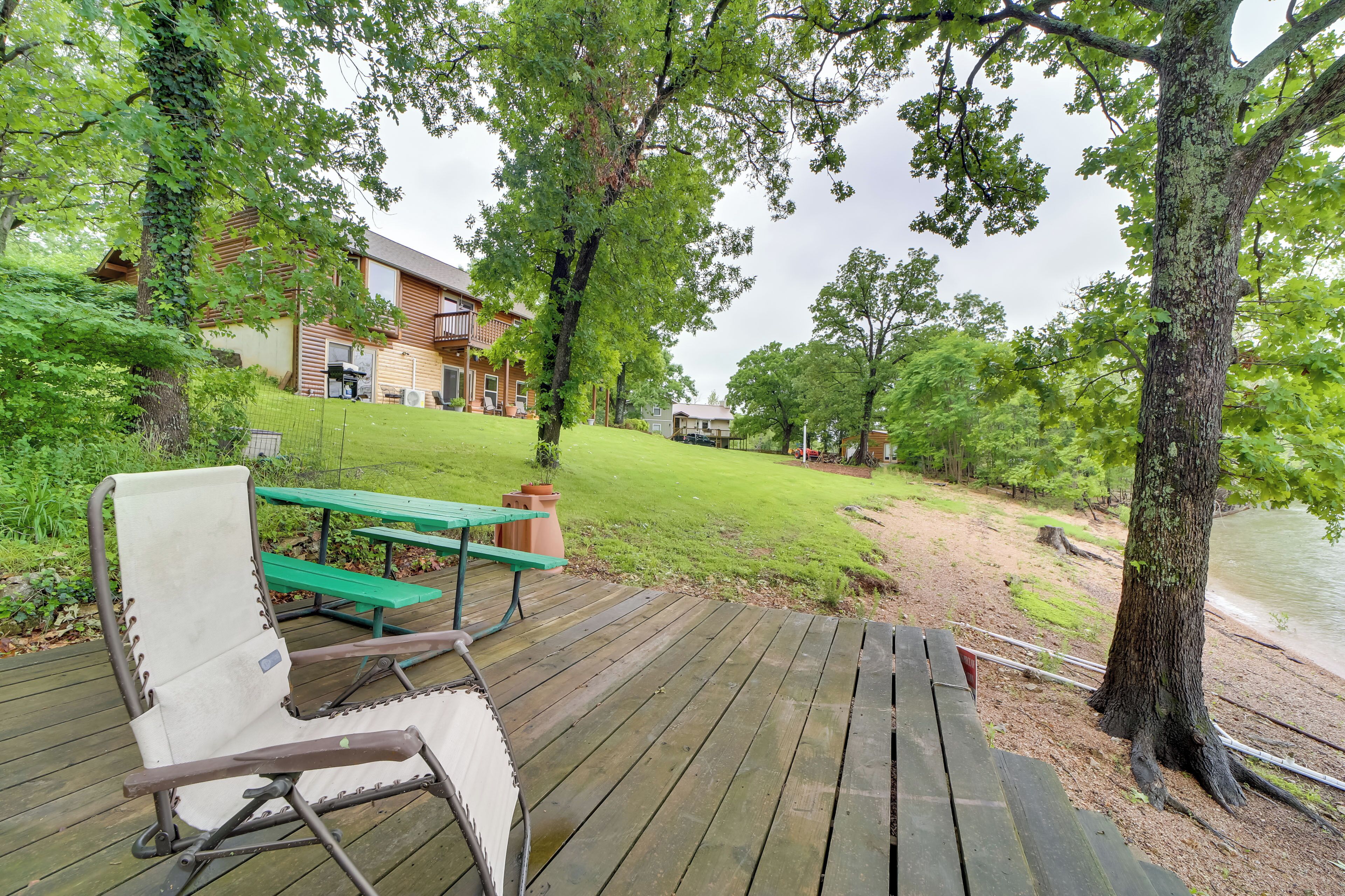 A wooden deck with a lounge chair and picnic table, overlooking a grassy area and a lake.