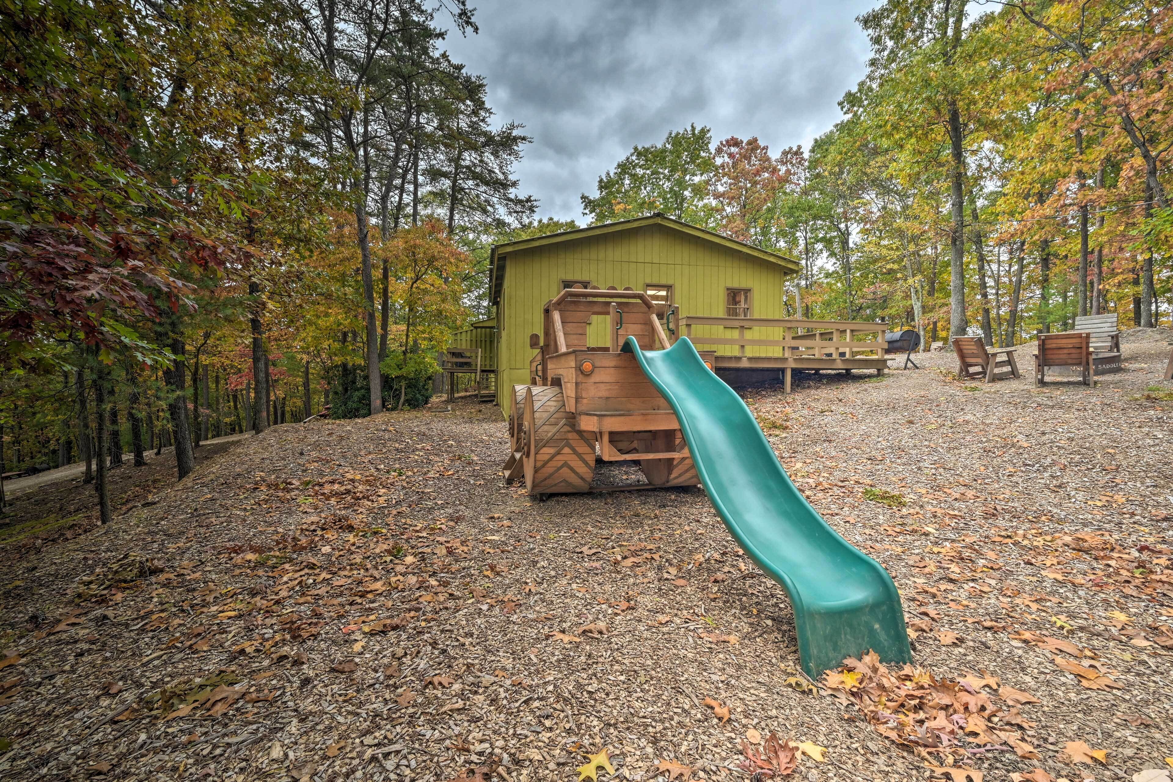 Un parque infantil con un tobogán y estructuras de juego de madera, rodeado de árboles y un edificio al fondo.