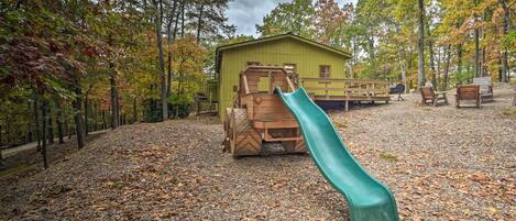Un parque infantil con un tobogán y estructuras de juego de madera, rodeado de árboles y un edificio al fondo.