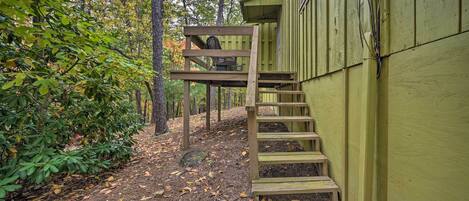 A wooden staircase leading to a deck with a chair, surrounded by trees and a green building.