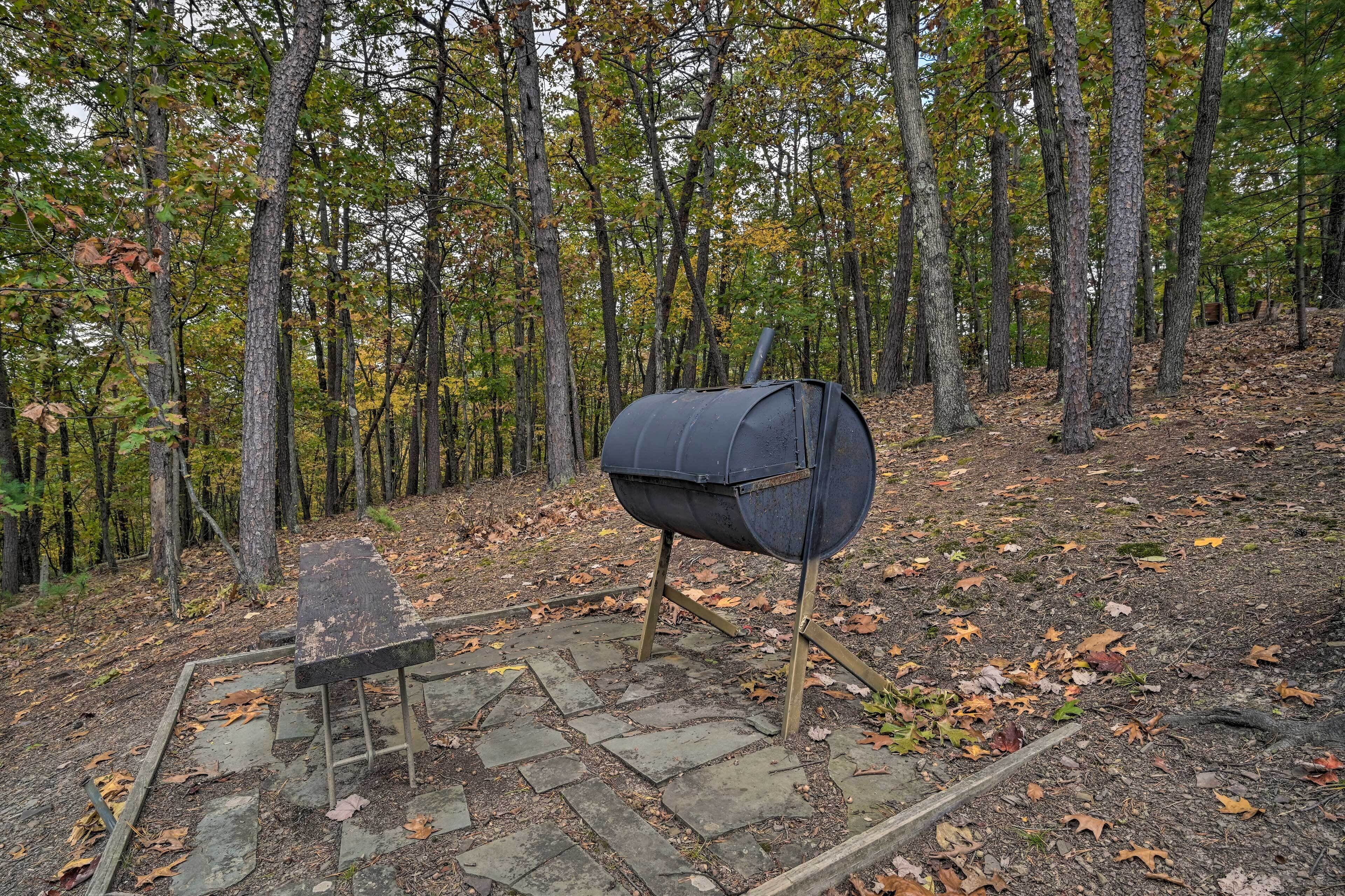 Un coin barbecue rustique aménagé à l’extérieur, dans une zone boisée, avec des marches en pierre et un banc.