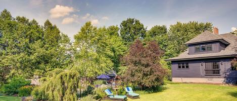 A house with a well-maintained lawn, a patio with two lounge chairs, and a large tree in the backyard.