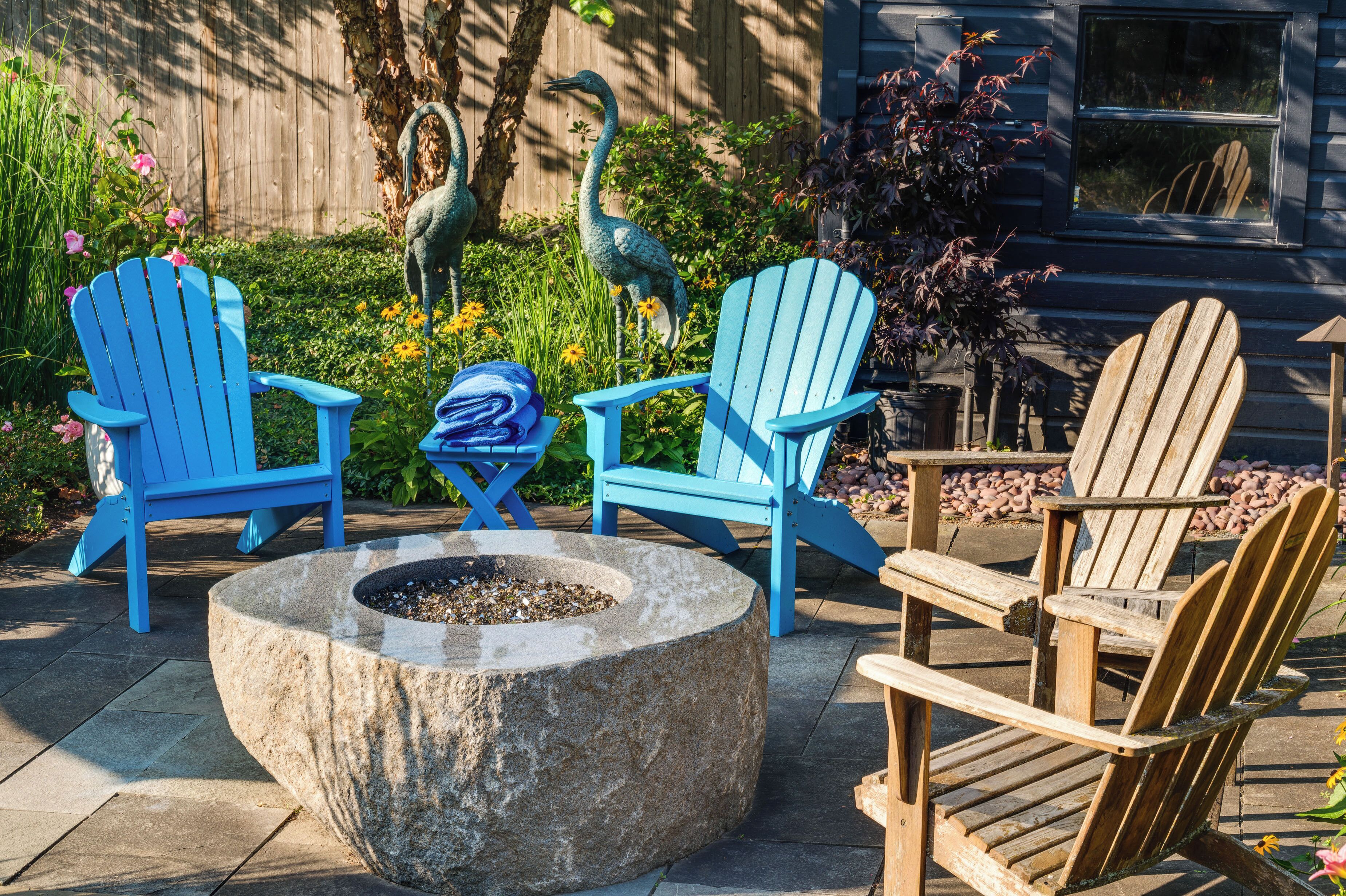 A garden with two blue Adirondack chairs, a stone fire pit, and a wooden chair.