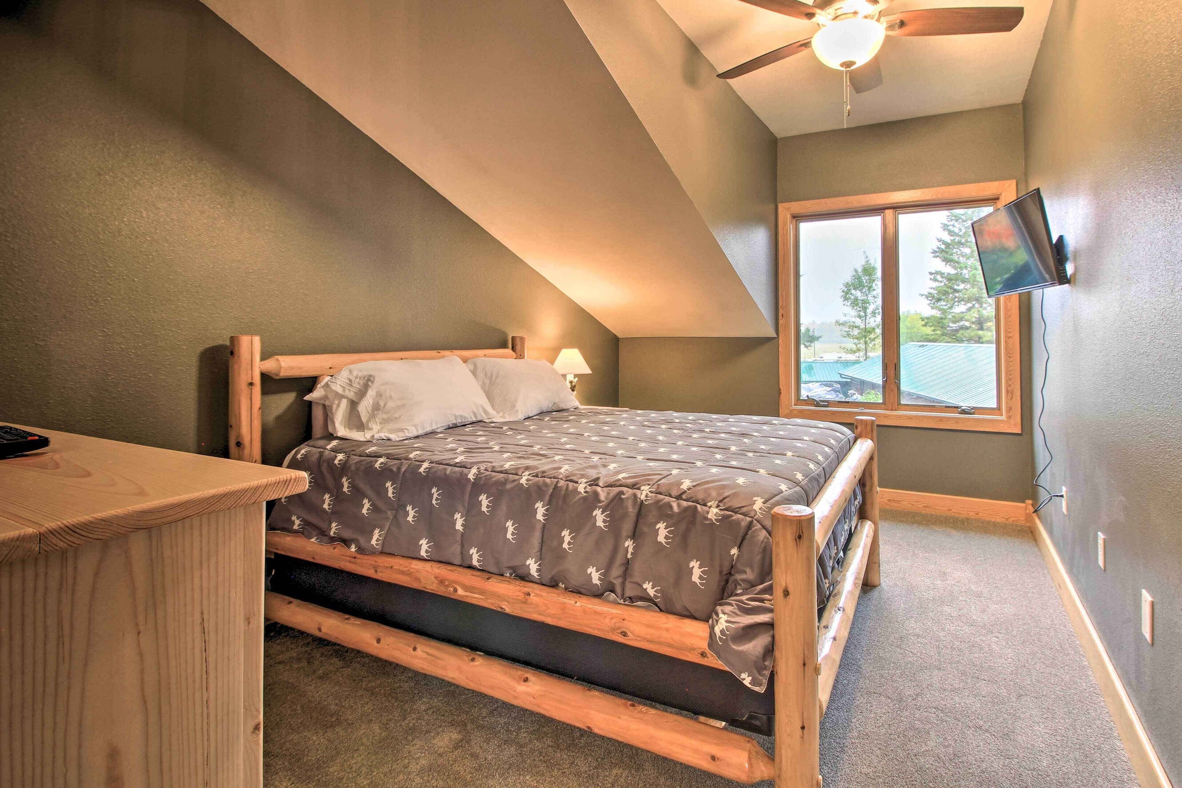 A bedroom with a wooden bed, a desk, a ceiling fan, a TV mounted on the wall, and a window with a view of the outdoors.