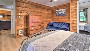 A room with wooden panel walls, a ceiling fan, a bed with patterned bedding, a blue sofa, and a dresser. - Game Room + Fire Pit: Weaverville Family Home! (Weaverville)