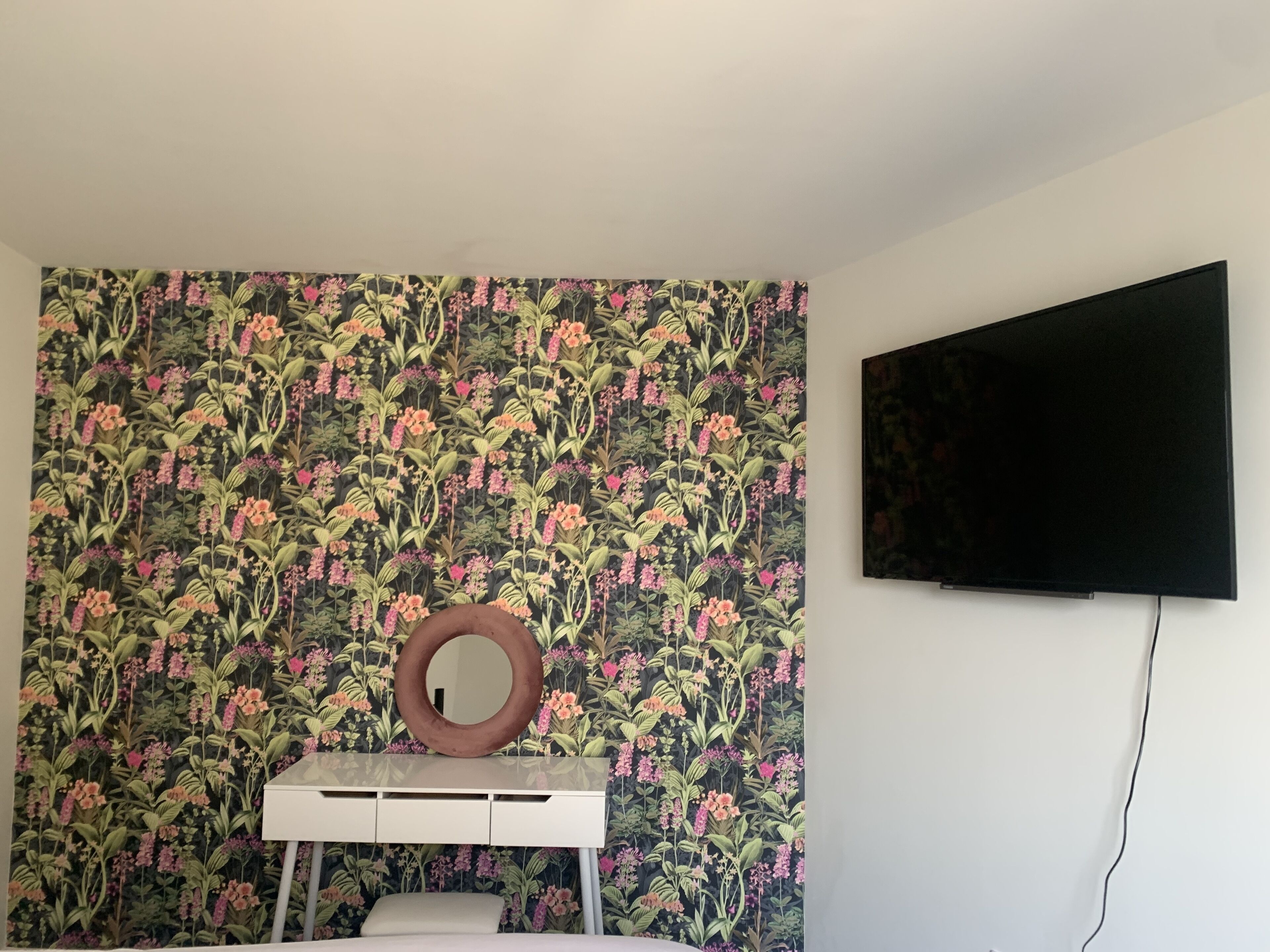 Una habitación con papel tapiz floral, un tocador blanco con espejo y un televisor montado.