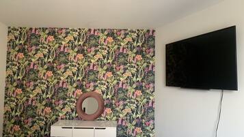 Una habitación con papel tapiz floral, un tocador blanco con espejo y un televisor montado.