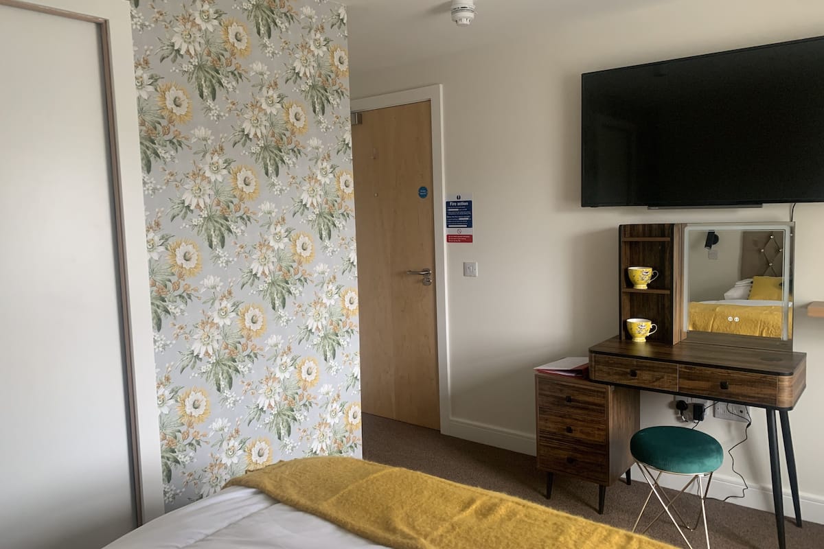 A hotel room with a bed, a desk with a chair, a television mounted on the wall, and a door with a fire action sign.