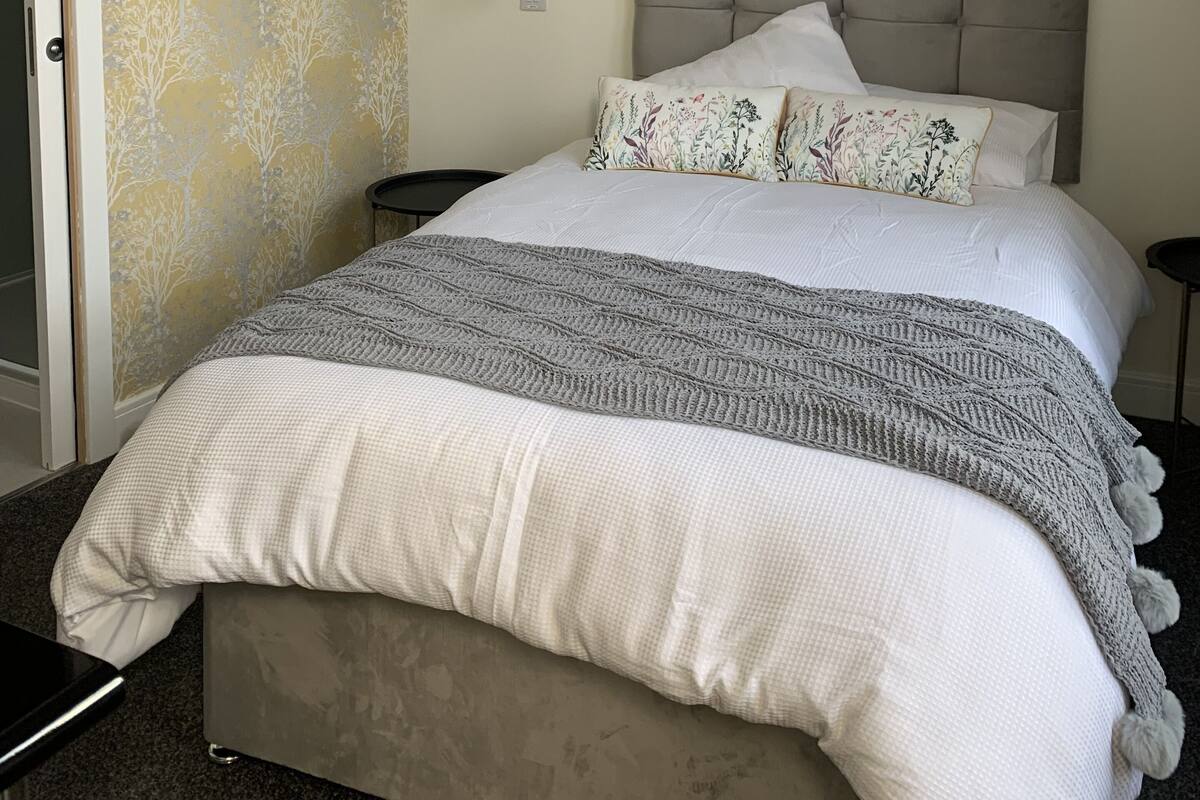 A bedroom with a bed, a headboard, a nightstand, a wall-mounted light, and a framed picture.