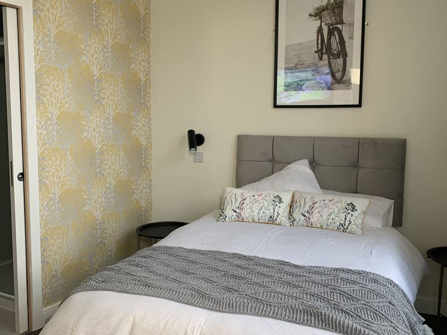 Een slaapkamer met een bed, een hoofdeinde, een nachtkastje, een wandlamp en een lijst met een afbeelding.