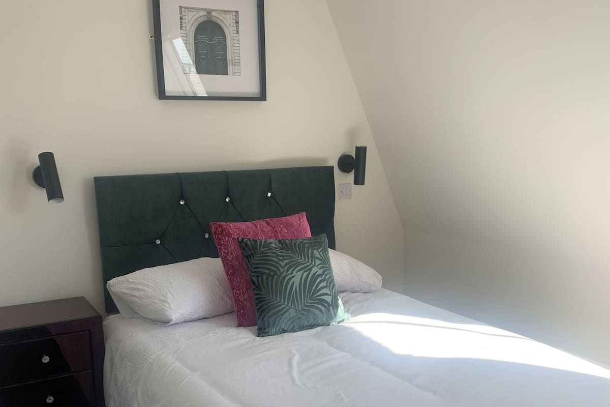 A neatly made bed with white bedding and a green headboard, a framed picture on the wall, and a dark nightstand with drawers.