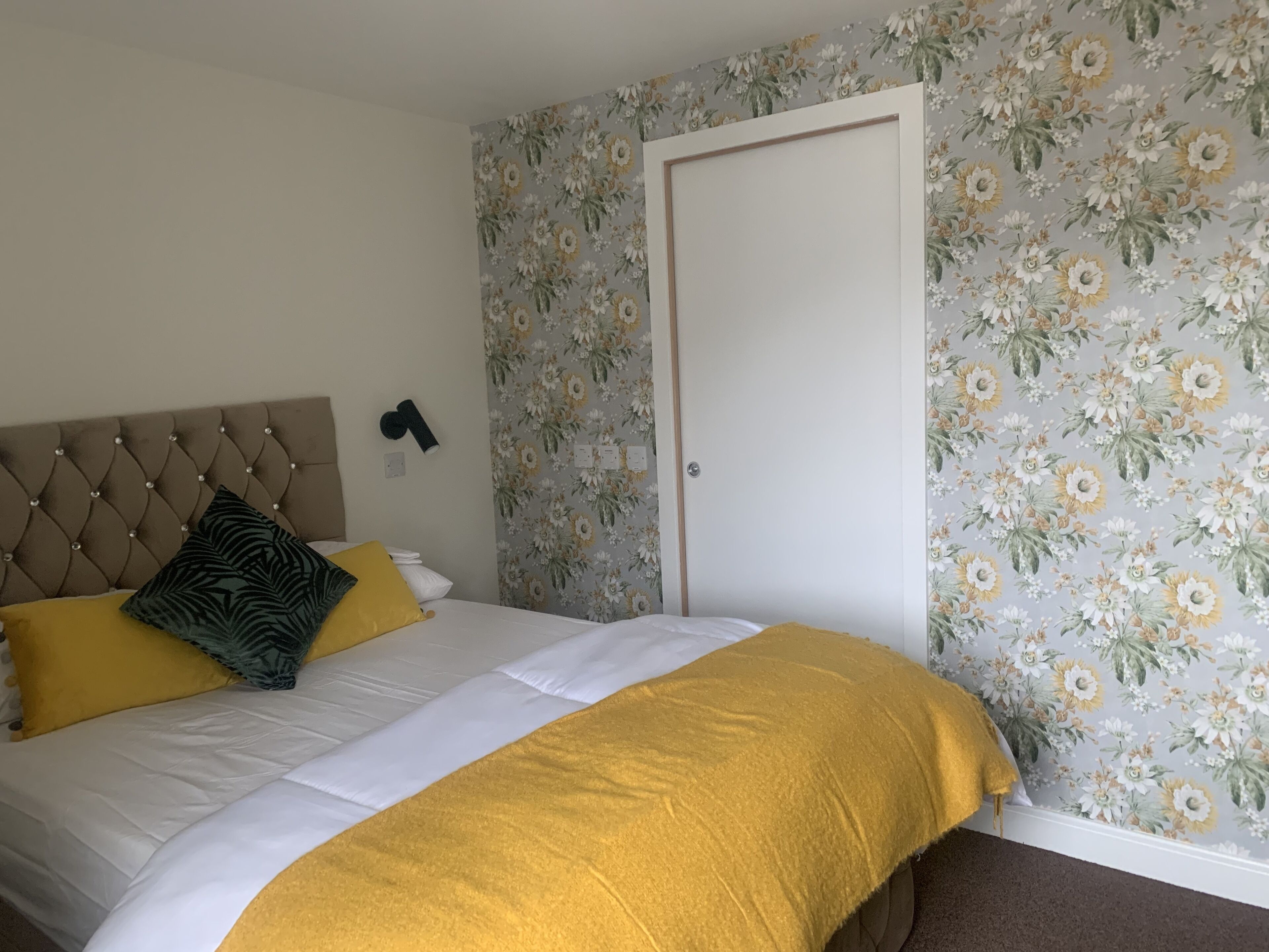 Ein Schlafzimmer mit einem gebürsteten, gepolsterten Kopfteil, einem Bett mit weißer und gelber Bettwäsche und floralem Wandpapier.
