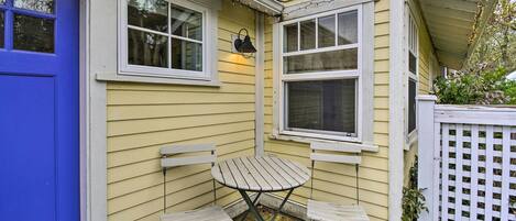 A small outdoor seating area with two white metal chairs and a table, adjacent to a yellow house with white trim and a blue door.