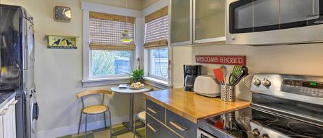 Una cocina con piso a cuadros, microondas, horno, cafetera y una pequeña mesa con una silla.