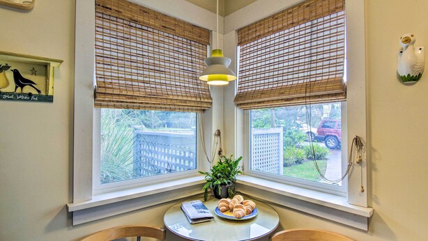 A corner room with two windows, a table with a plate of croissants, a potted plant, and a wall-mounted bird sculpture.