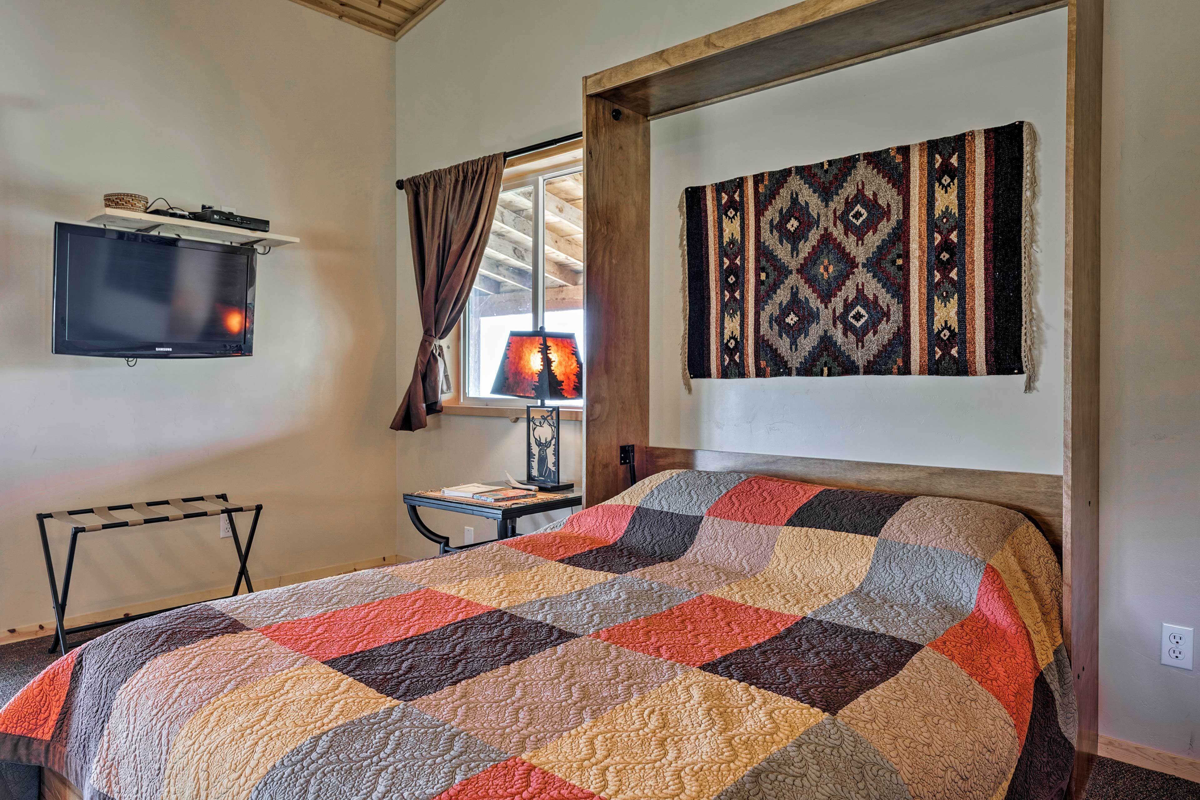 A bedroom with a large bed, a mounted TV, a desk with a lamp, and a window with curtains.