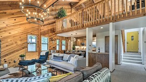 A spacious living room with a high ceiling, wooden beams, a balcony with a railing, a sofa set, a glass coffee table, and a chandelier. - Hike, Fish, & Relax: Family Arnold Cabin (Arnold)