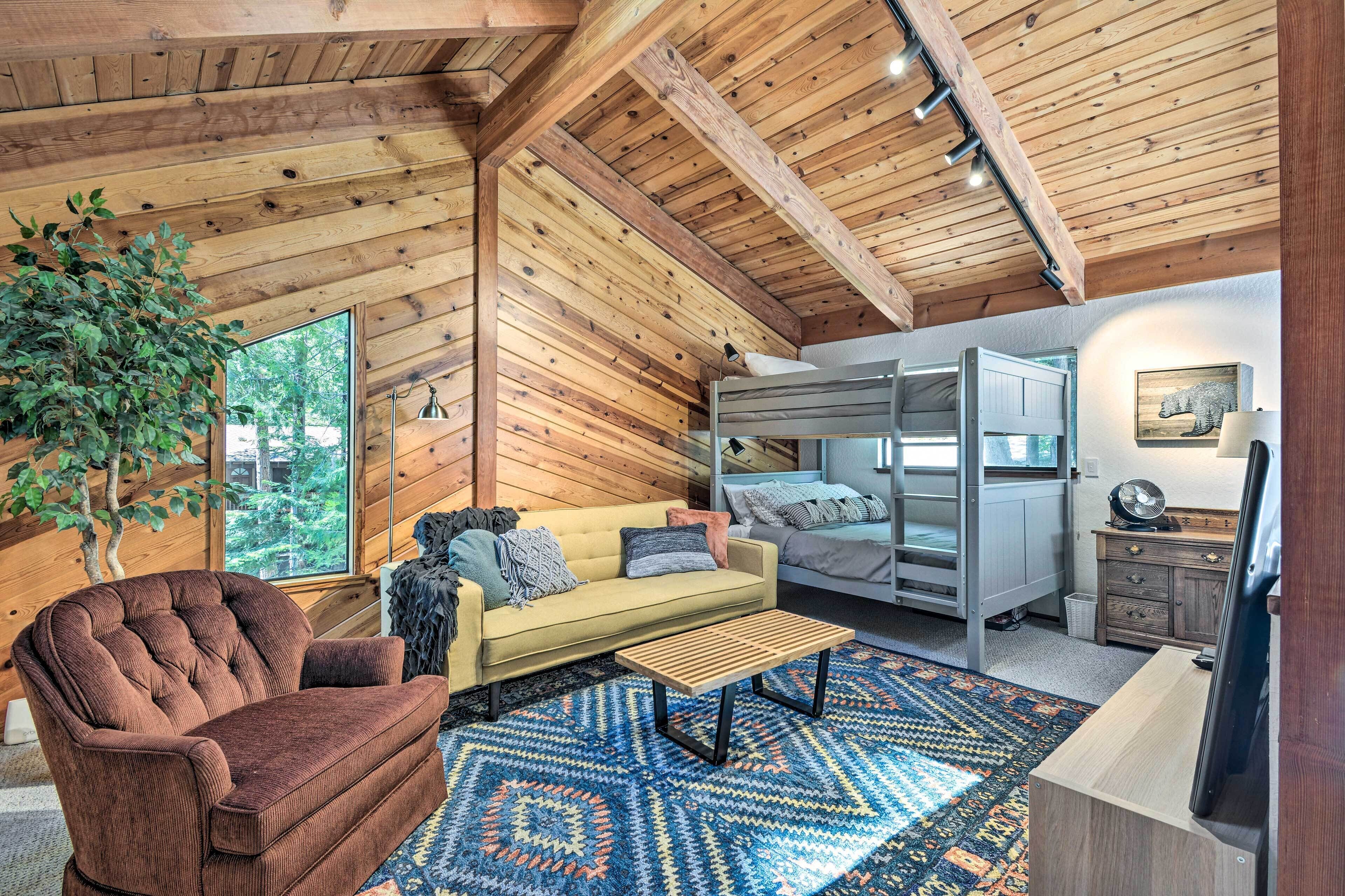 A cozy cabin interior with a wooden ceiling, a sofa, a coffee table, a bunk bed, and a large window offering a view of trees.
