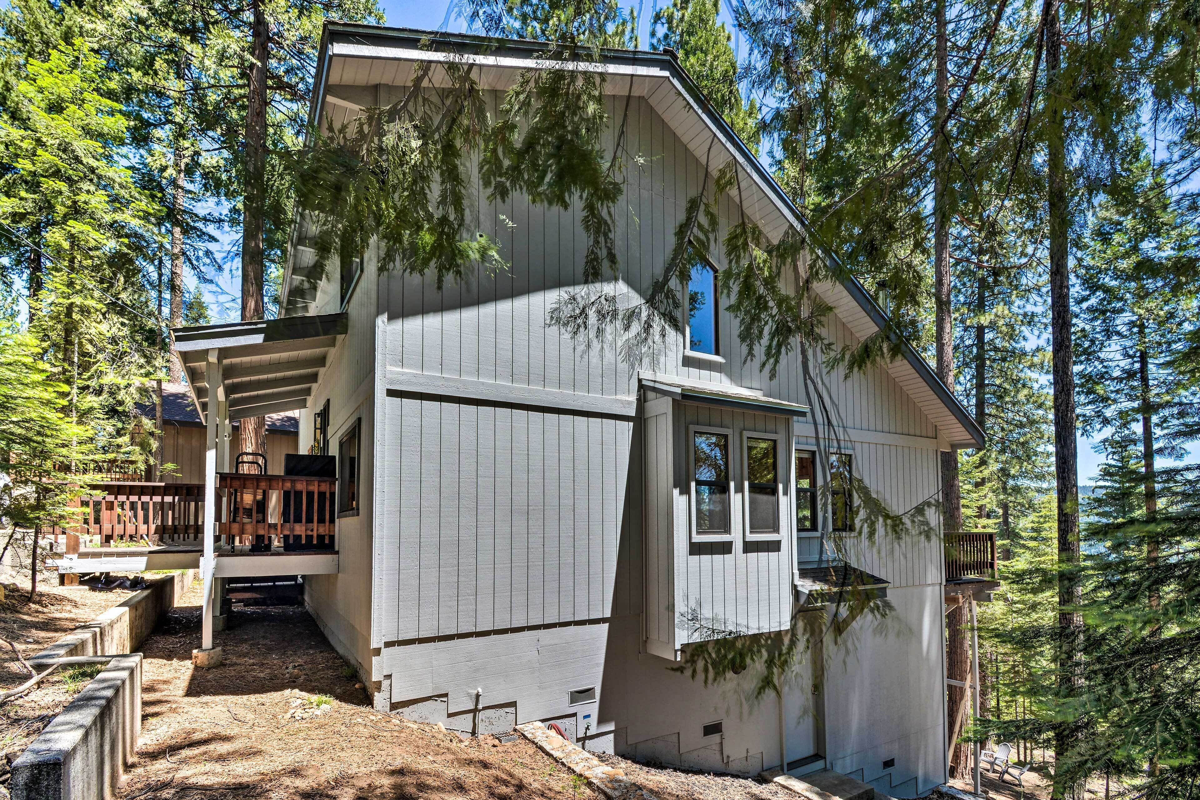 A two-story cabin with a deck and a large window, surrounded by tall pine trees.