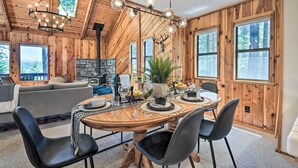 A wooden interior with a dining table, chairs, a chandelier, a fireplace, and large windows. - Hike, Fish, & Relax: Family Arnold Cabin (Arnold)