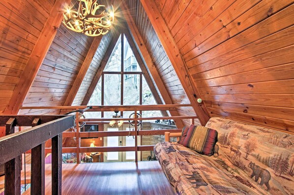 A cozy attic bedroom with a wooden sloped ceiling, a large window, a wooden staircase, and a sofa with patterned cushions.