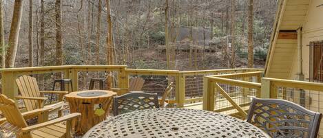 A wooden deck with Adirondack chairs, a metal table, and a view of a forested area.