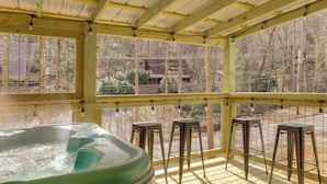 A hot tub area with a wooden deck, metal stools, and a view of a forested area through large windows. - A-frame Gatlinburg Cabin w/ Deck & Private Hot Tub (Gatlinburg)