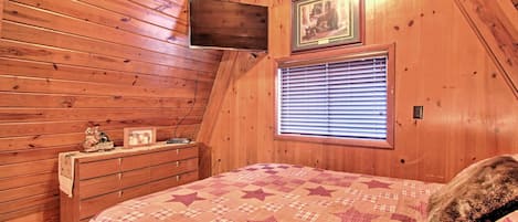 A cozy bedroom with wooden walls, a bed with a star-patterned quilt, a dresser, a flat-screen TV, and a window with blinds.