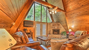 A cozy cabin interior with a fireplace, leather seating, and a large window overlooking greenery. - A-frame Gatlinburg Cabin w/ Deck & Private Hot Tub (Gatlinburg)