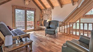 Un interior rústico con pared de piedra, vigas de madera, un sillón de cuero y vistas a través de amplios ventanales.