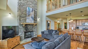A spacious living area with a stone fireplace, a large flat-screen TV, a grey sectional sofa, and a wooden dining set. - Getaway Chalet in Mars Hill w/ Hot Tub & Views! (Mars Hill)